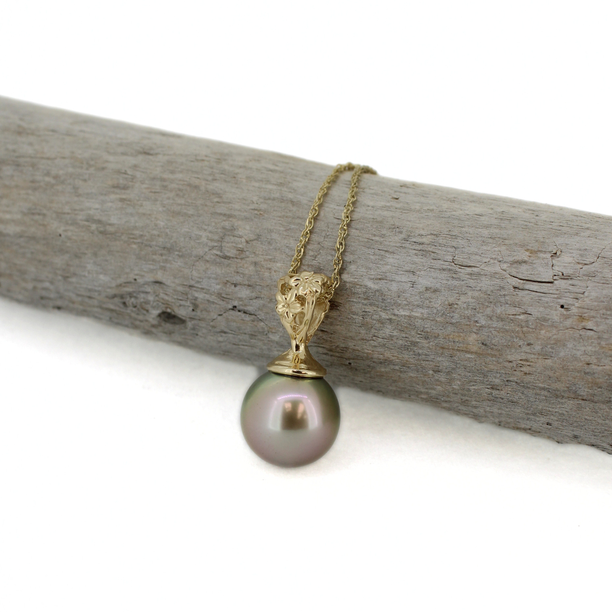 A gold pendant necklace featuring a single round, iridescent gray-green pearl suspended from an ornate gold cap on a delicate gold chain, displayed on a piece of weathered wood against a white background.