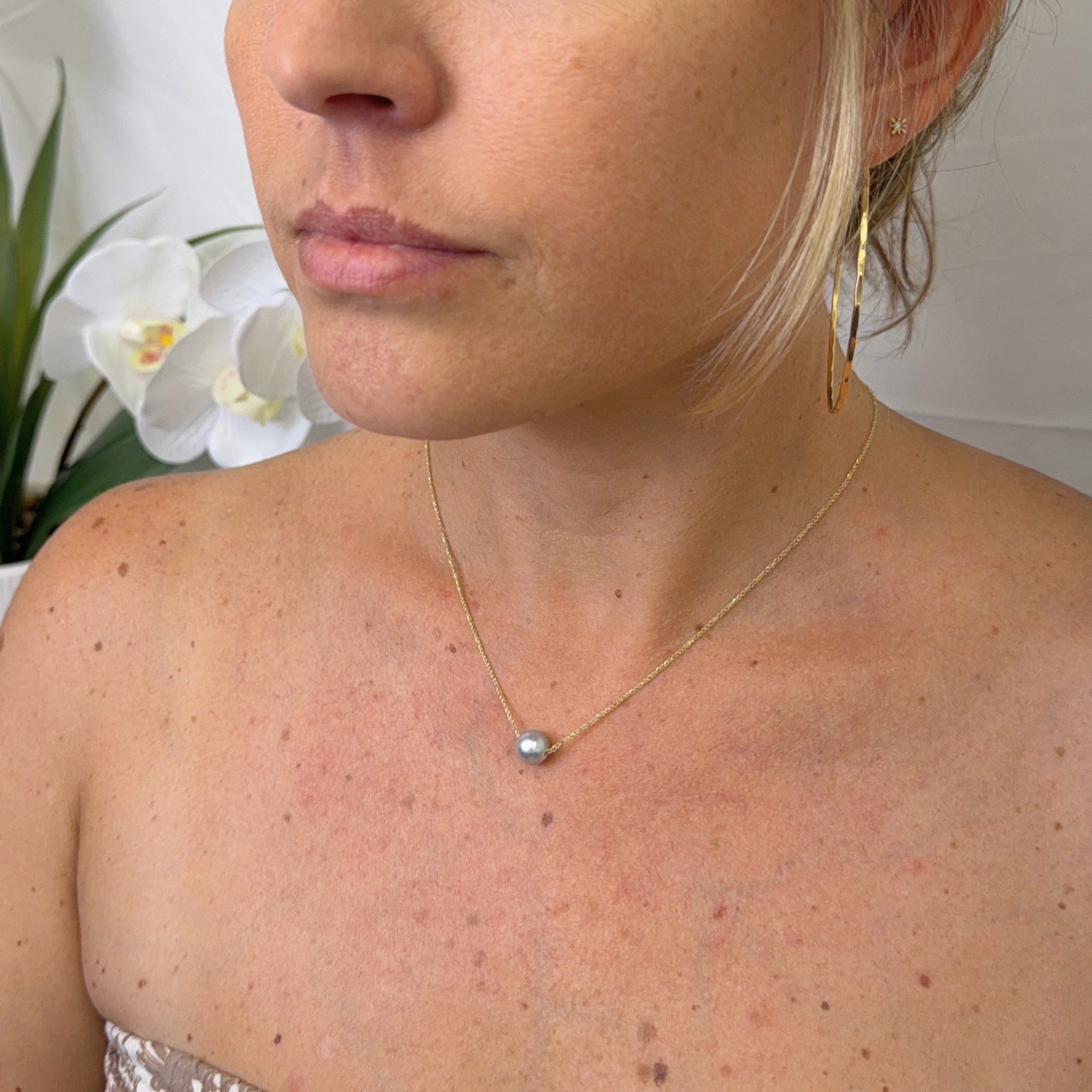 A close-up lifestyle shot of a woman wearing a delicate gold chain necklace with a small, round, light blue-grey akoya pearl. The background is softly blurred, showing white orchids on the left.