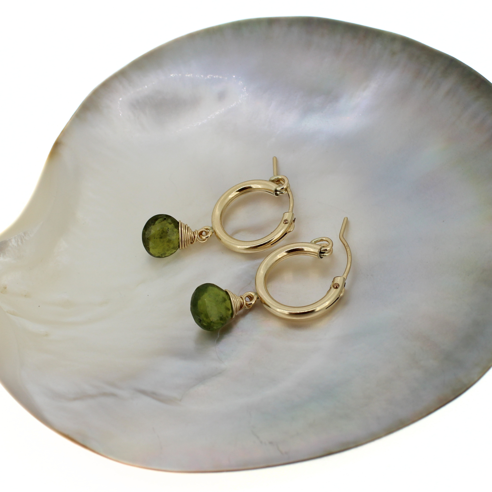 A pair of gold hoop earrings with small green idocrase gemstones, displayed on a white mother-of-pearl shell against a white background.