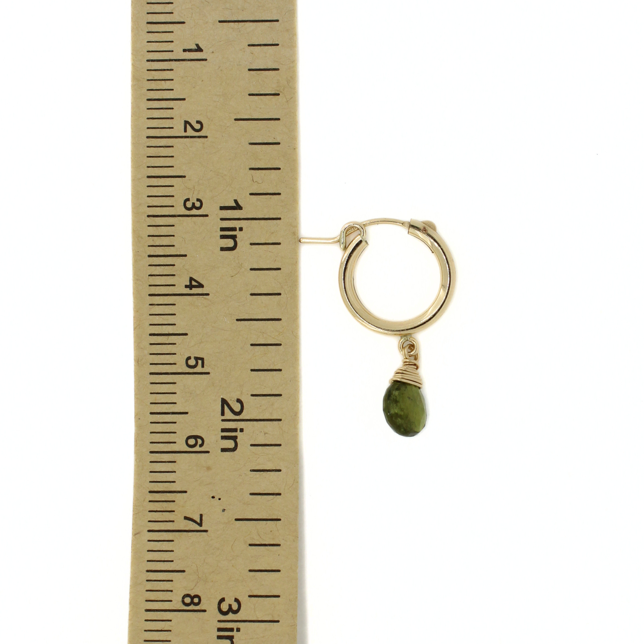 A single gold hoop earring with a small green teardrop stone dangle, shown next to a paper ruler for scale on a white background.