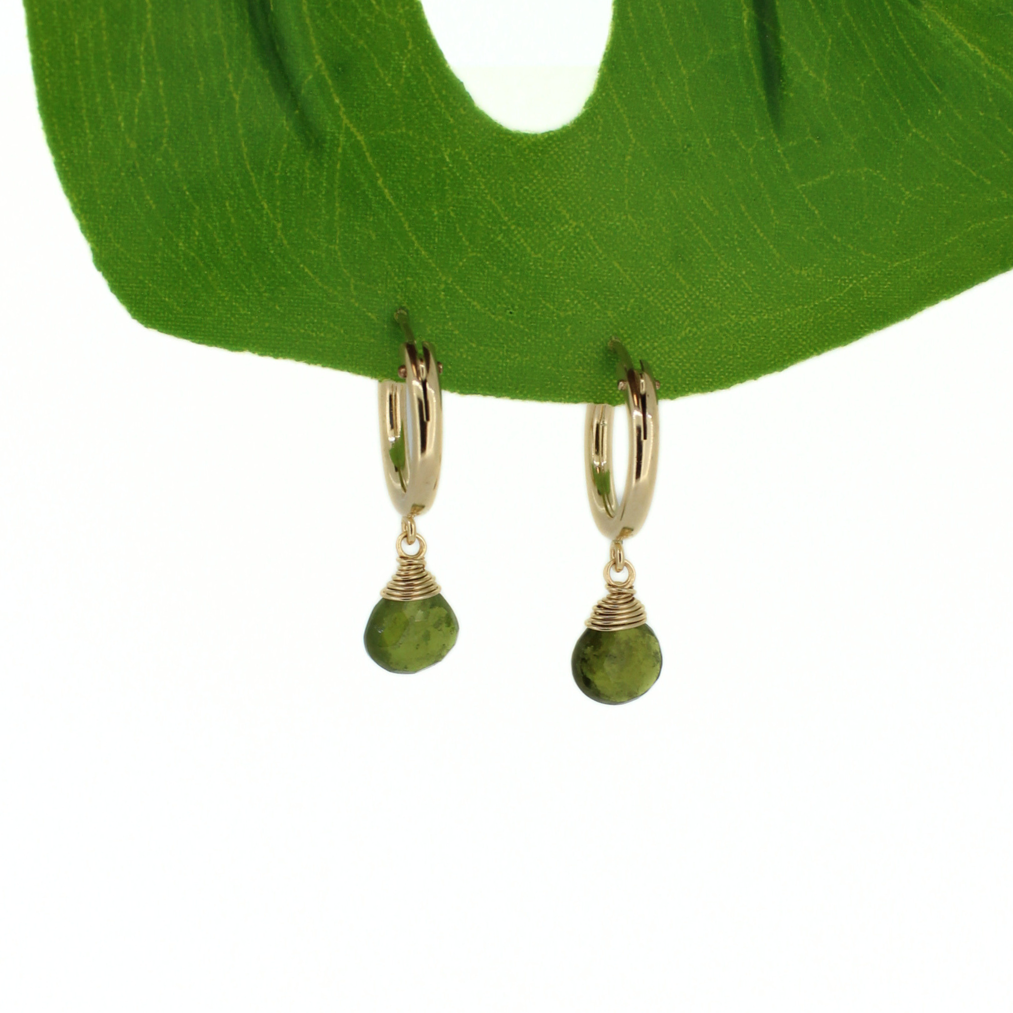 A pair of gold huggie hoop earrings with green teardrop gemstones wrapped in wire, hanging from a green leaf display on a white background.