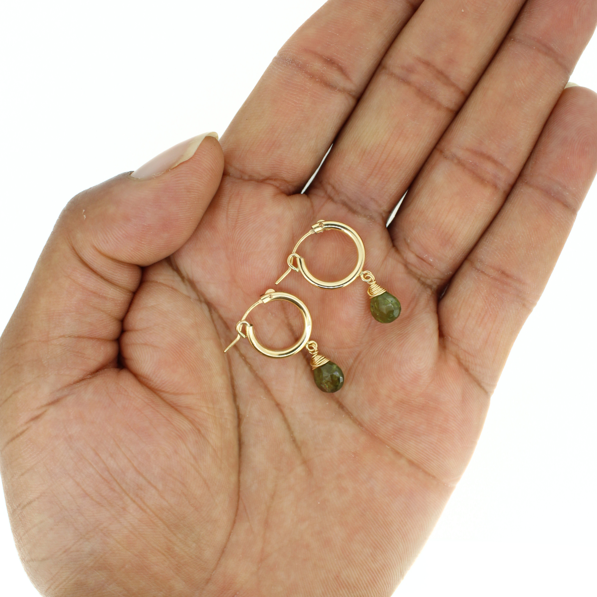 A pair of small gold hoop earrings with dangling green bead accents, displayed on an open hand against a white background.