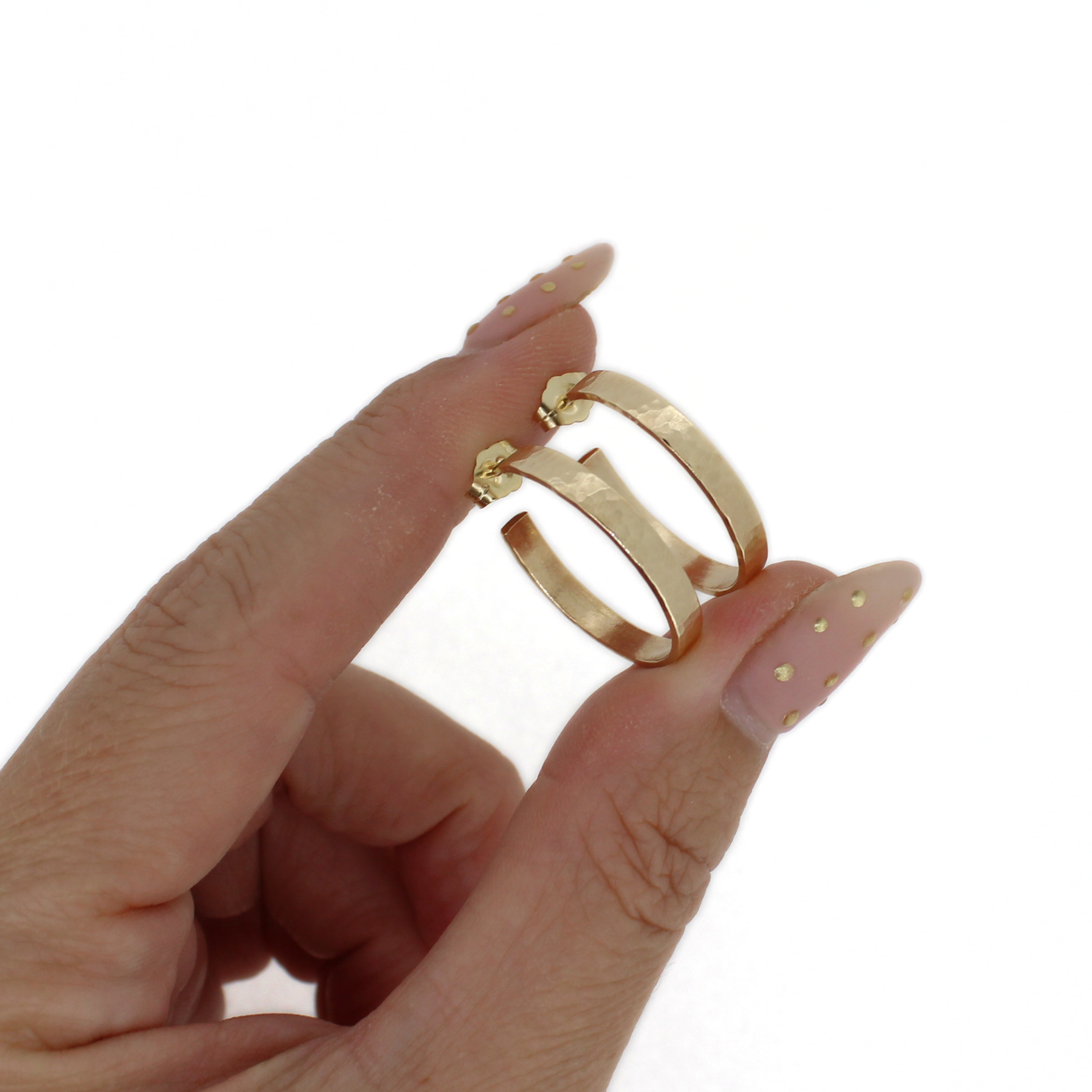 A close-up of a hand with manicured nails holding a pair of gold hoop earrings against a plain white background.
