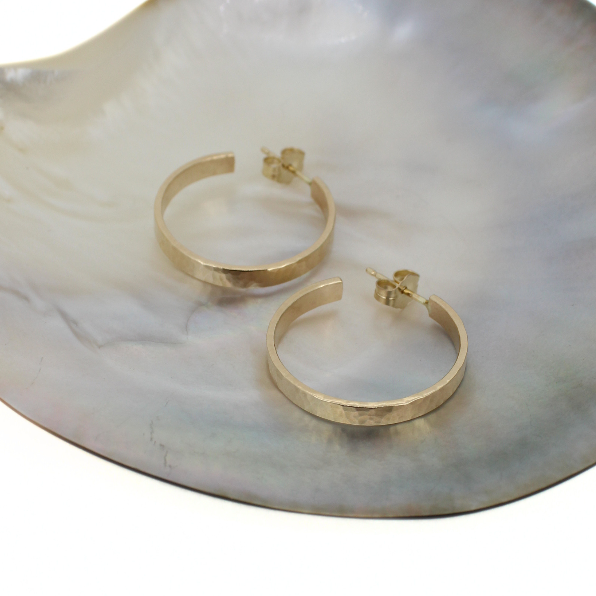 A pair of hammered gold hoop earrings displayed on a white and gray iridescent shell dish against a white background.