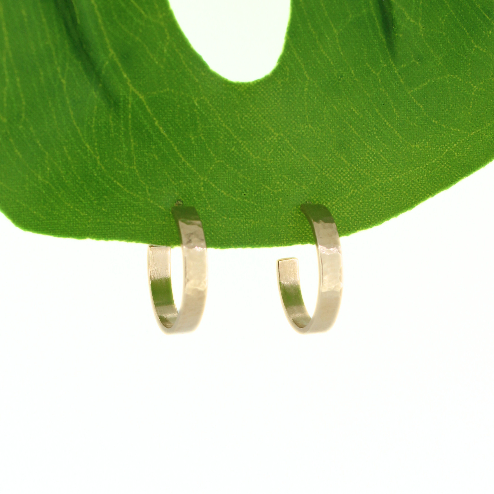 A pair of small open hoop earrings in gold, hanging from a bright green leaf against a white background. Handmade in Hawaii by Brianne & Co.