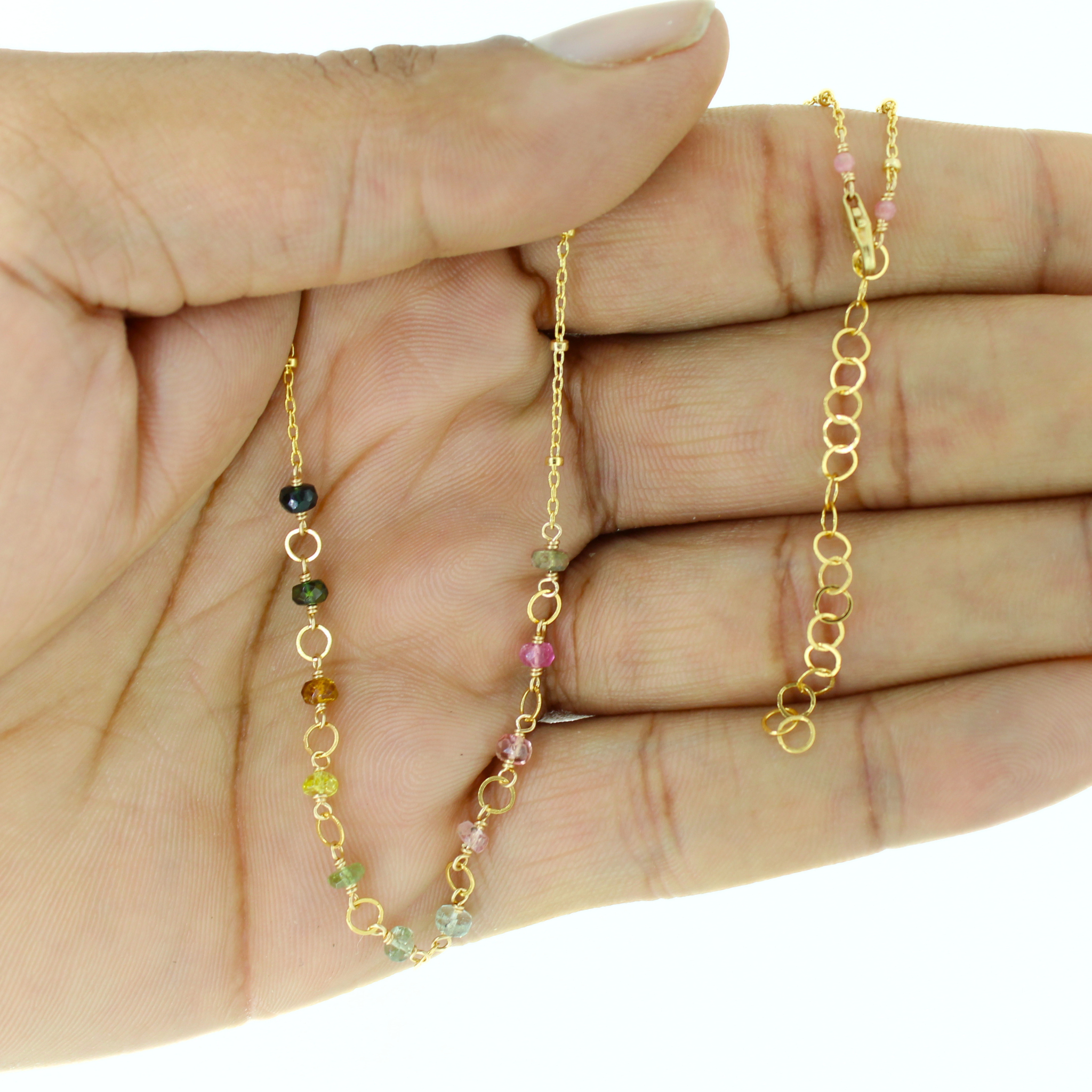 A delicate gold chain necklace with small multicolored gemstone beads and openwork gold links, held in a person's hand against a white background.