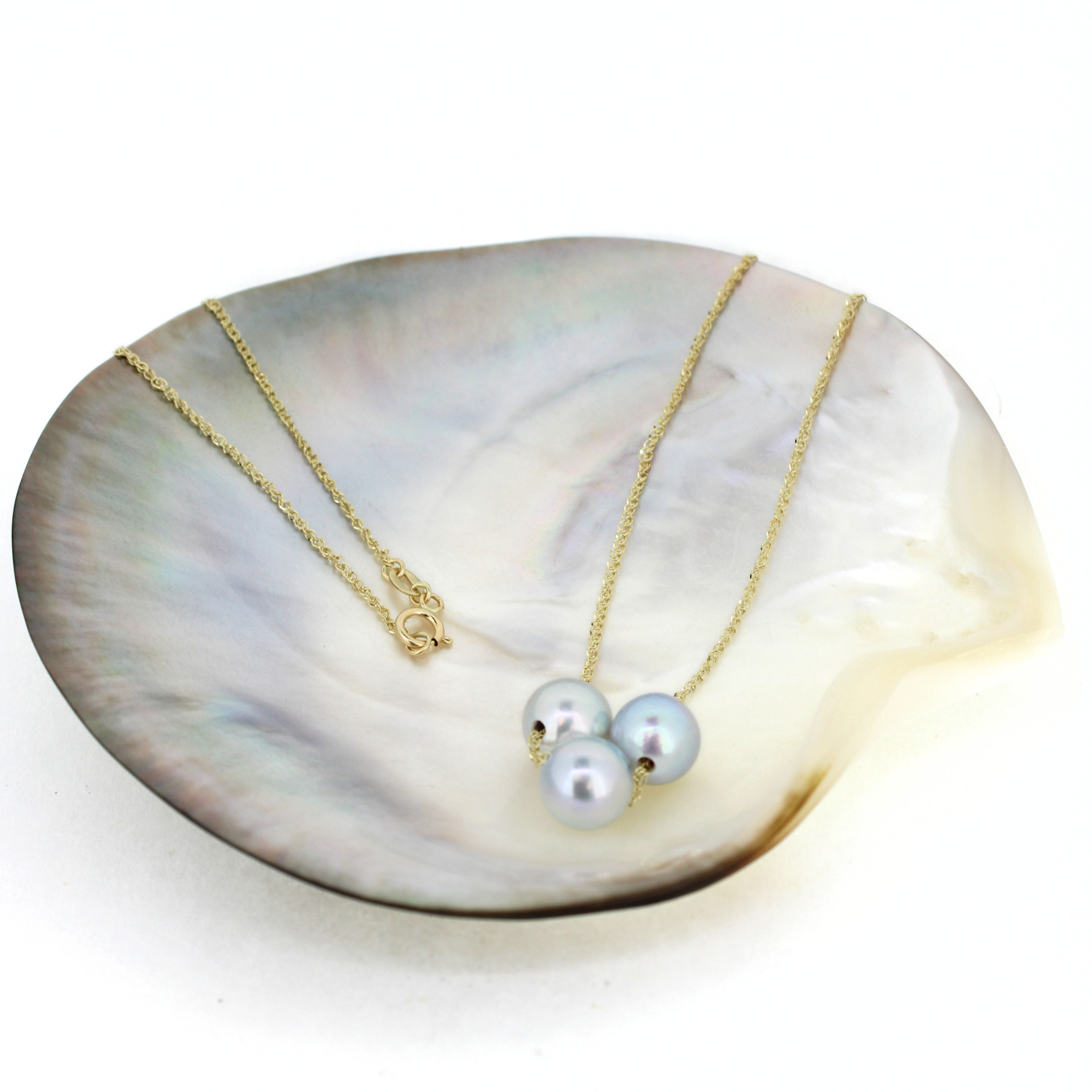 A delicate gold necklace with three light blue-gray pearls, displayed on a large iridescent mother-of-pearl shell dish against a white background.