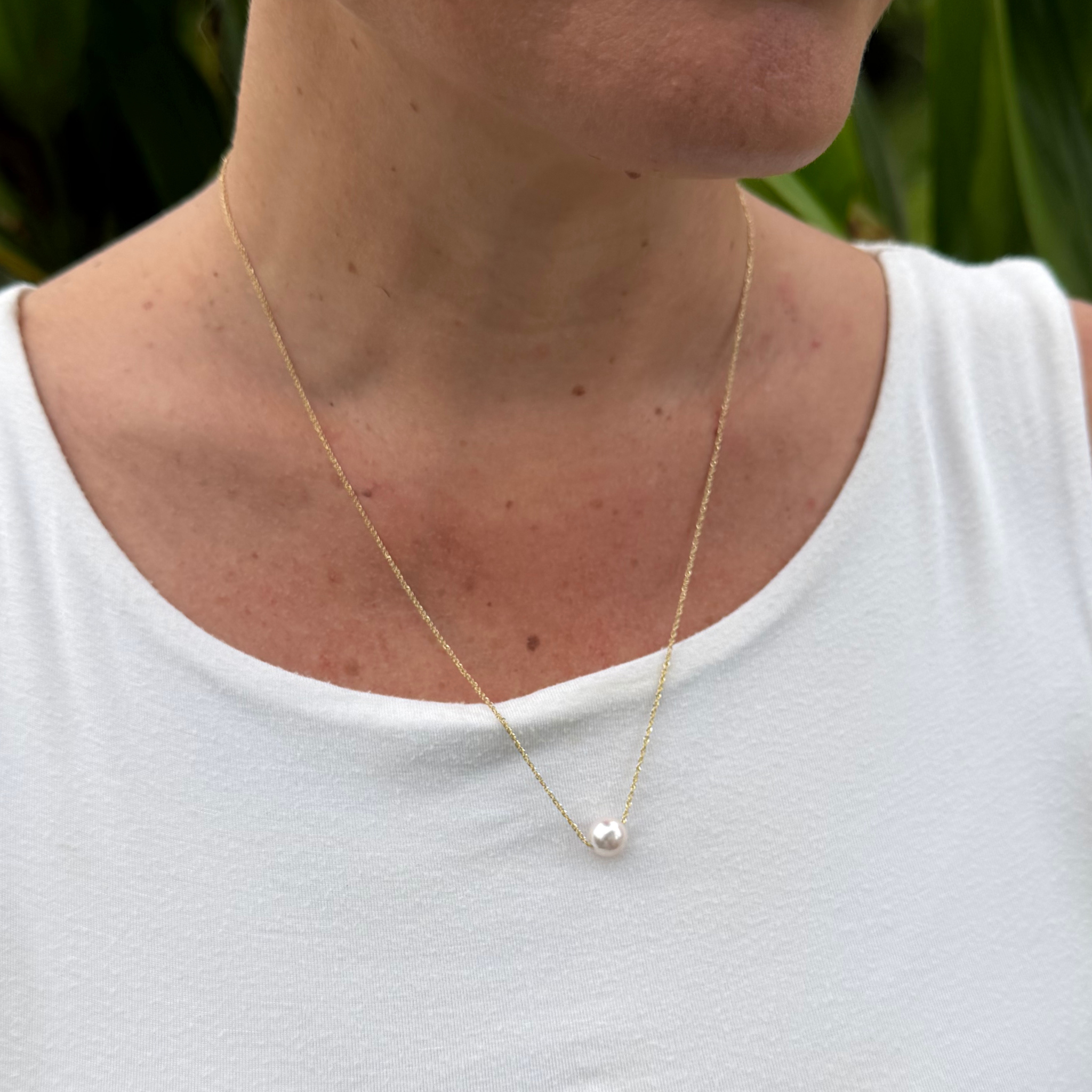 A close-up of a person wearing a delicate gold chain necklace with a small white akoya pearl, paired with a white sleeveless top.