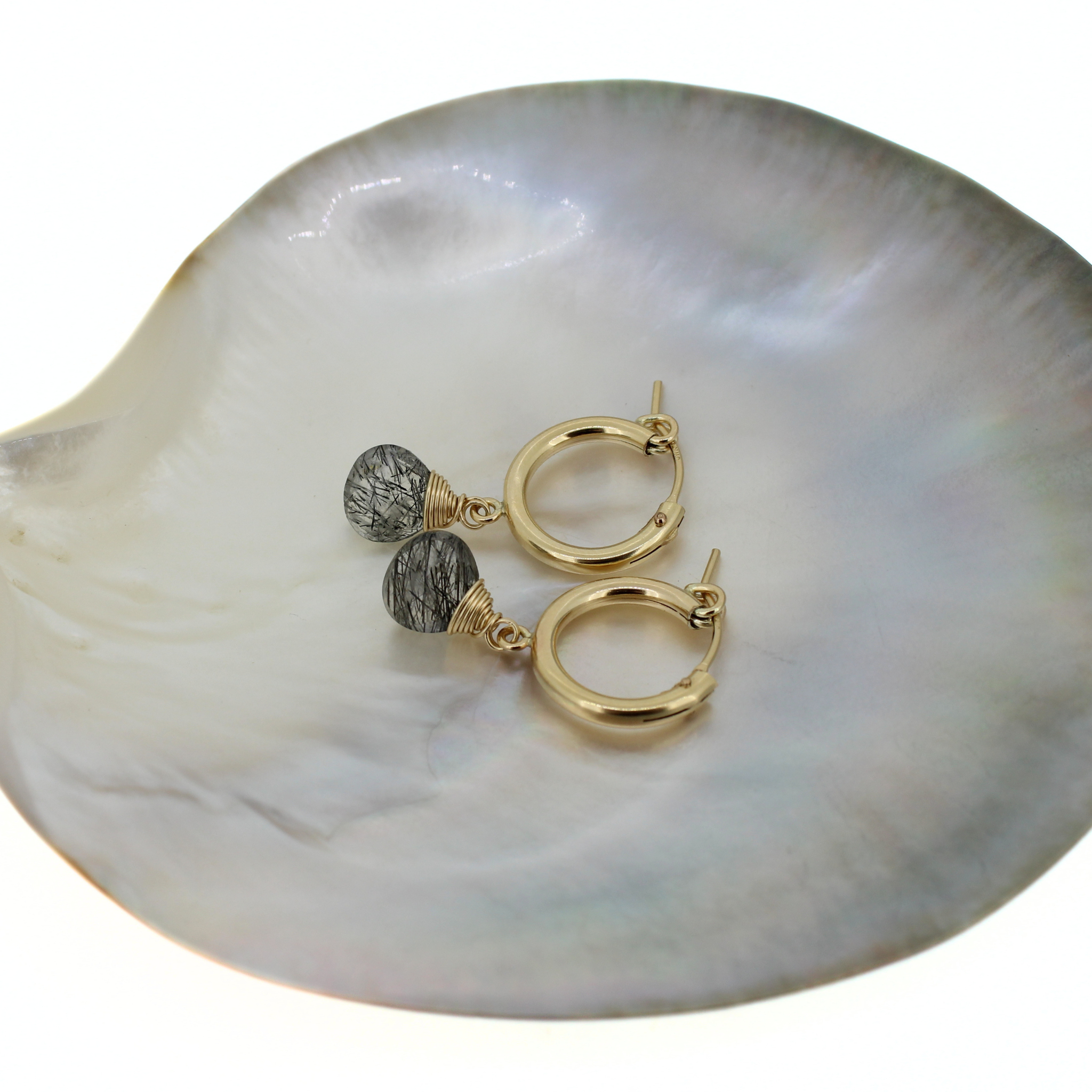 A pair of gold hoop earrings with faceted gray and black rutile quartz beads, displayed on a white iridescent shell dish against a white background.