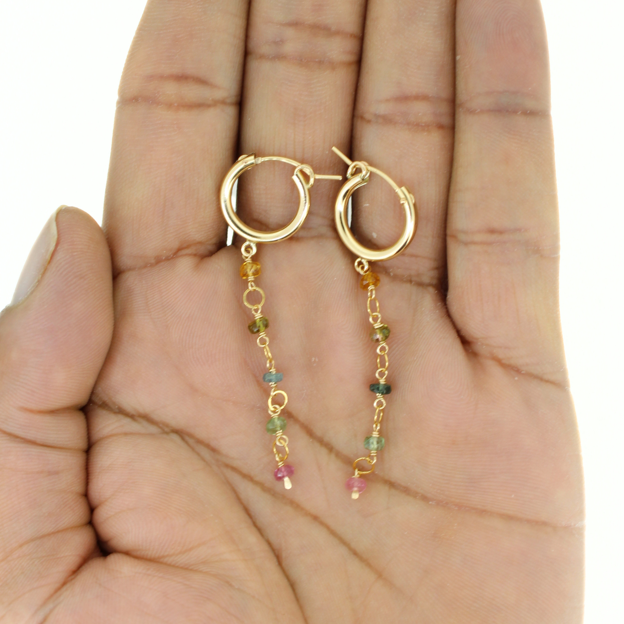 A pair of gold hoop dangle earrings with small multicolored beads, held in the palm of a hand.