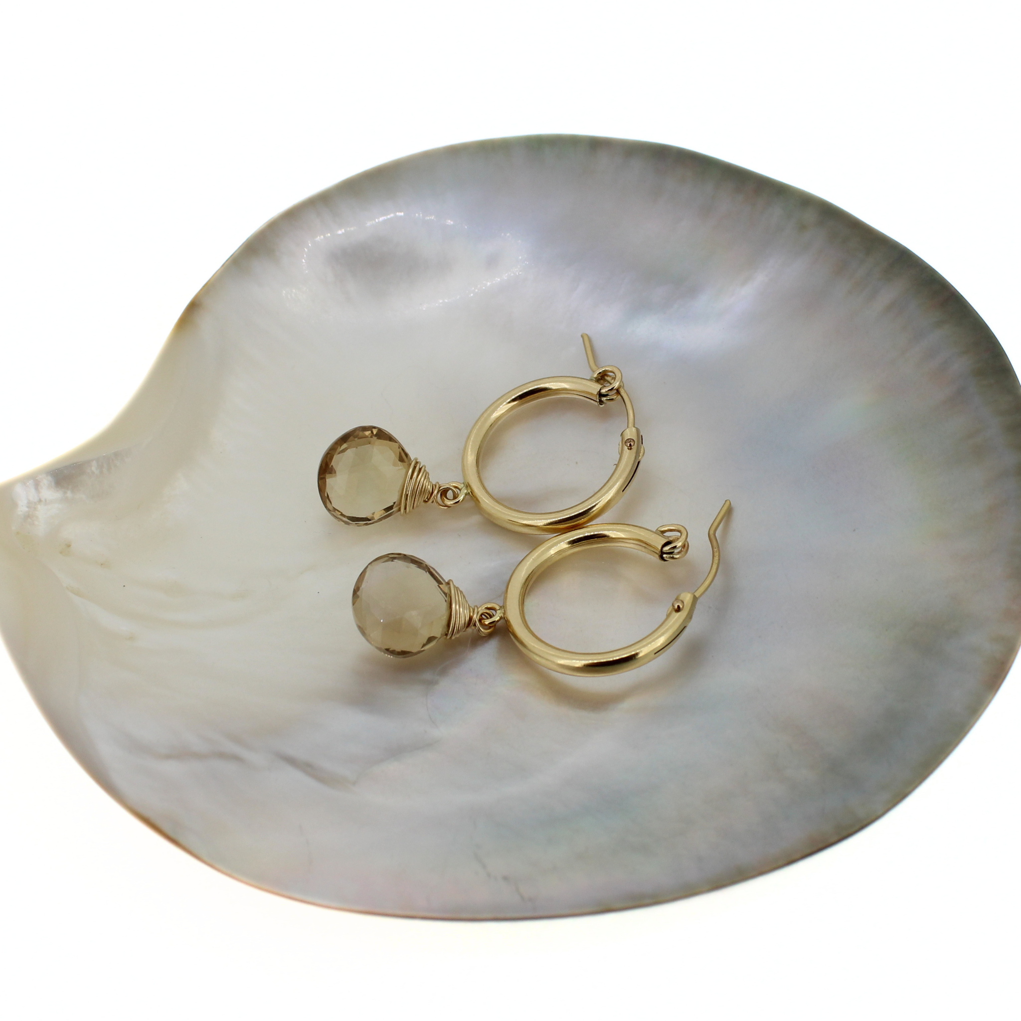 A pair of gold hoop earrings with small tear drop topaz gemstones, displayed on a white iridescent shell dish against a white background.
