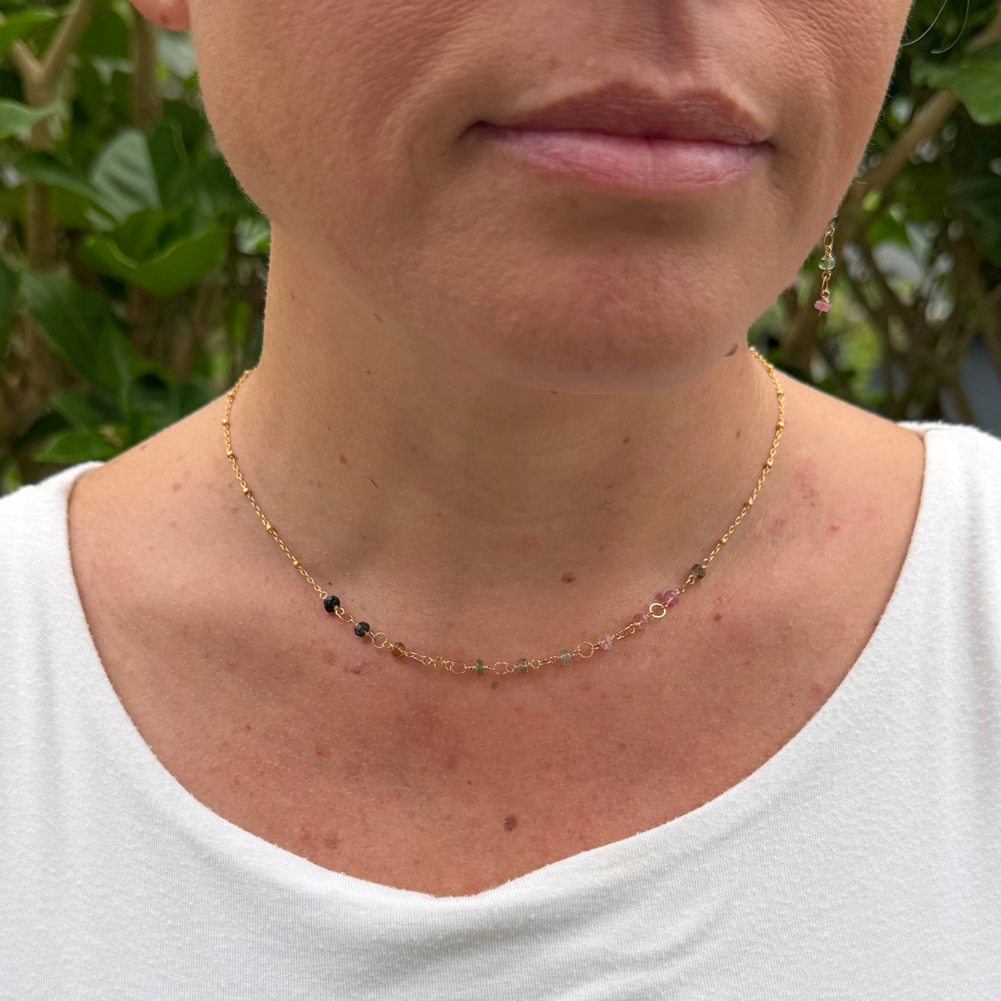 A close-up of a woman wearing a delicate gold chain necklace with small multicolored tourmaline gemstone accents, paired with a white top and an outdoor green background.