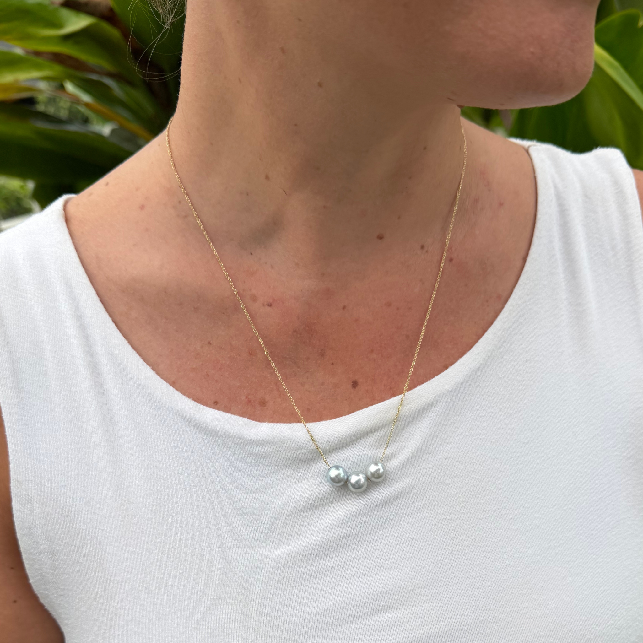 A close-up of a person wearing a delicate gold chain necklace with three genuine akoya pearls, styled with a white sleeveless top and a blurred green plant background.