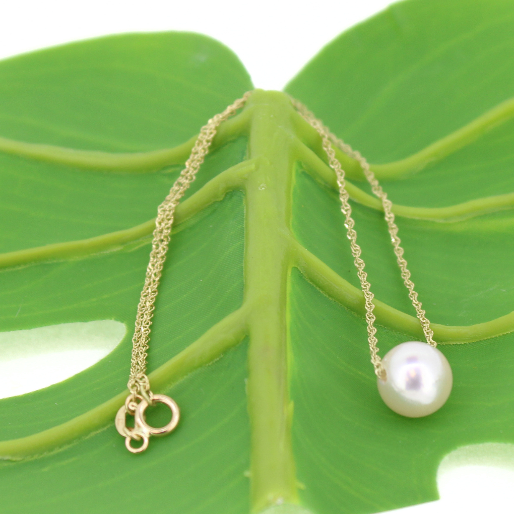 A delicate gold chain necklace with a single lustrous white pearl pendant, displayed on a large green leaf against a white background.