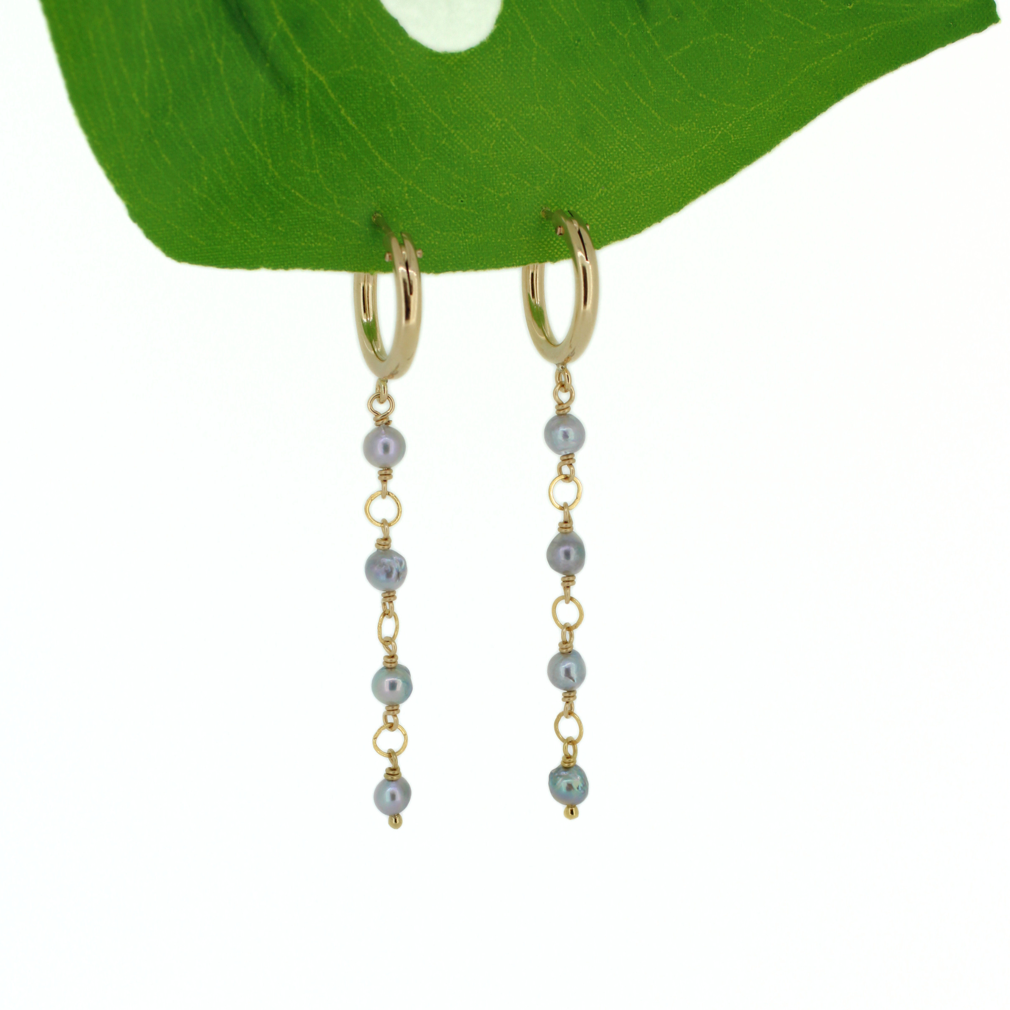 A pair of gold huggie hoop dangle earrings with small gray-blue akoya pearls, hanging from a green leaf against a white background.