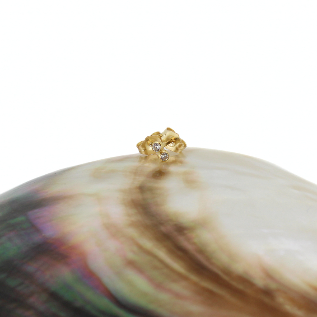A small gold stud earring set with clear cubic zirconias set on the edge of a large iridescent abalone shell, photographed against a solid white background.