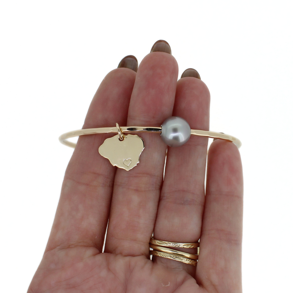A gold bangle bracelet with a small gold Kauai-shaped charm and a gray pearl, held on a person's hand against a white background.