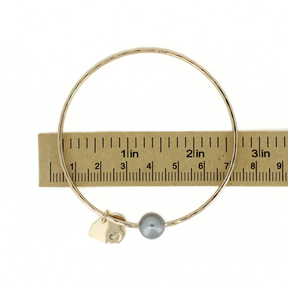 A gold bangle bracelet with a small gray pearl and a gold Kauai charm, shown next to a wooden ruler for scale on a white background.