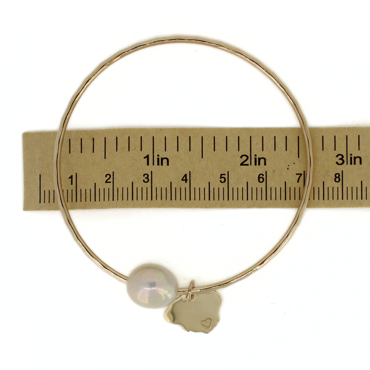 A gold wire bangle bracelet with a white pearl and a small gold charm, shown next to a paper ruler for scale on a white background.