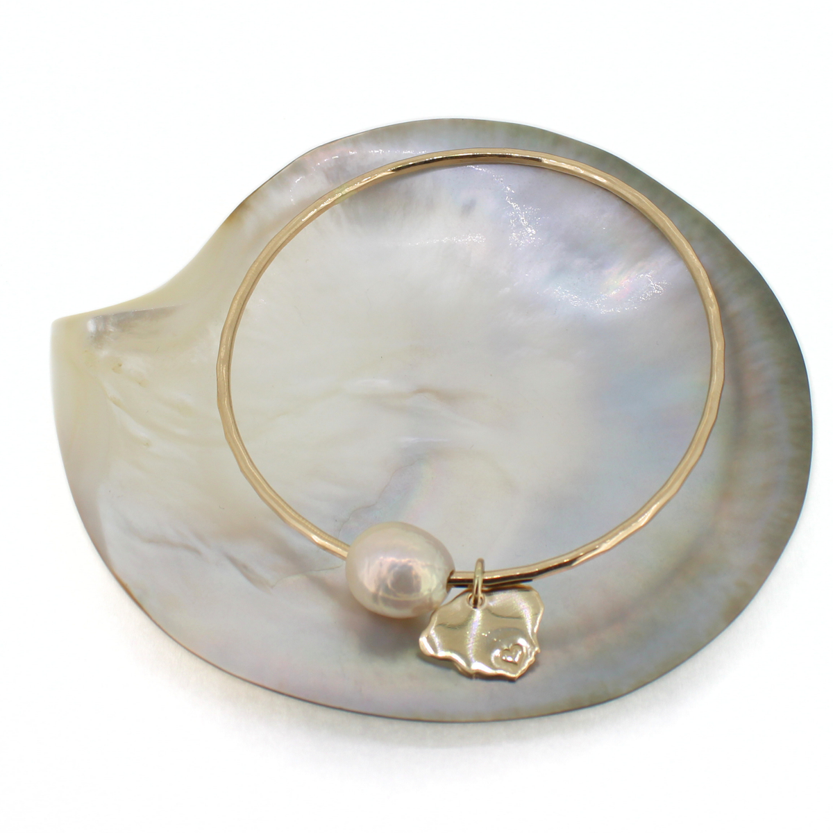 A delicate gold bangle bracelet with a baroque pearl and a small Kauai charm, displayed on a large iridescent mother-of-pearl shell against a white background.