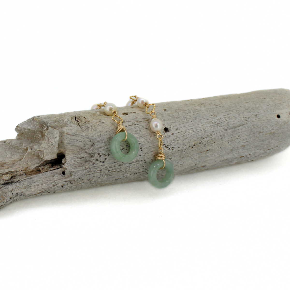 A pair of gold-toned earrings with green jadeite ring pendants and small white pearls, displayed on a piece of weathered driftwood against a white background.