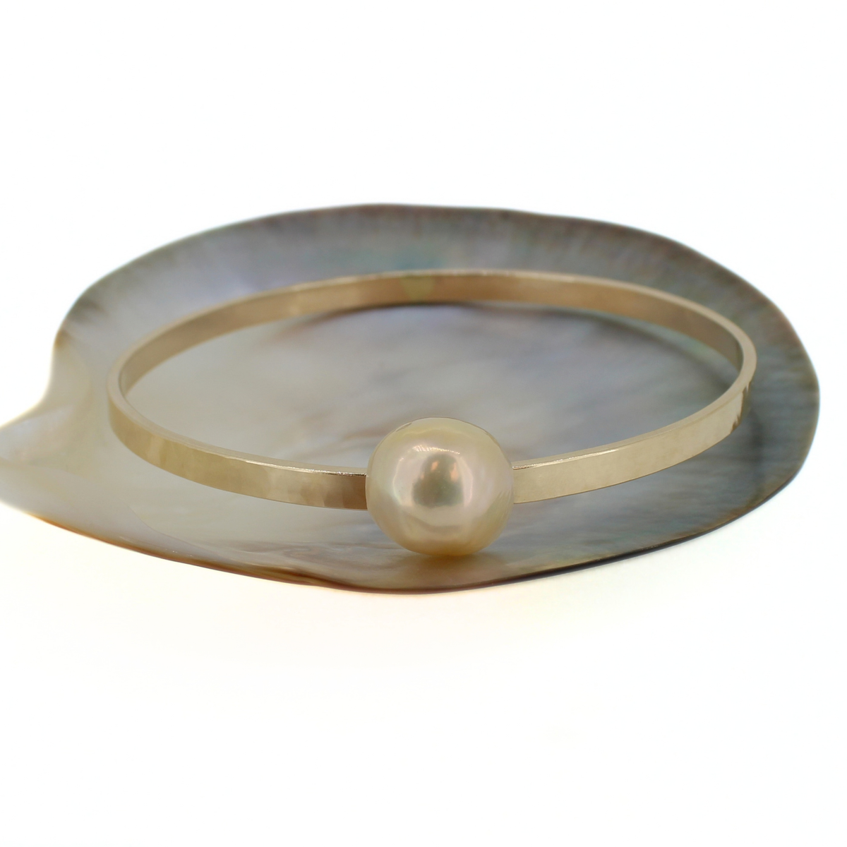 A minimalist hammered gold bangle bracelet with a single baroque pearl, displayed on a mother-of-pearl shell stand against a white background.