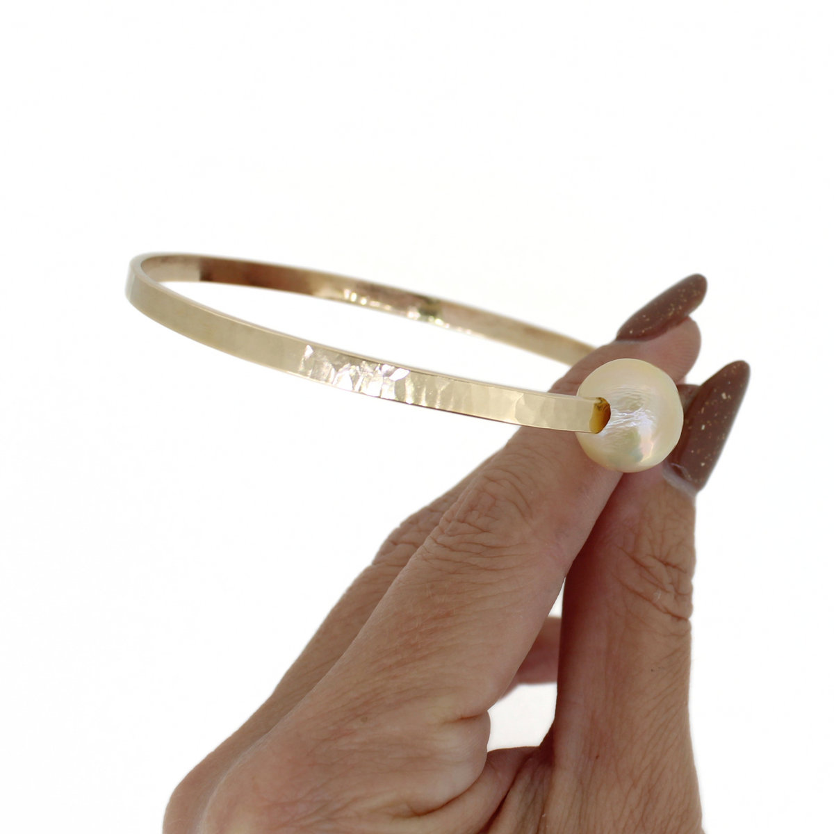 A gold bangle bracelet with a textured hammered finish and a round off-white pearl, held by a hand with brown manicured nails against a white background.