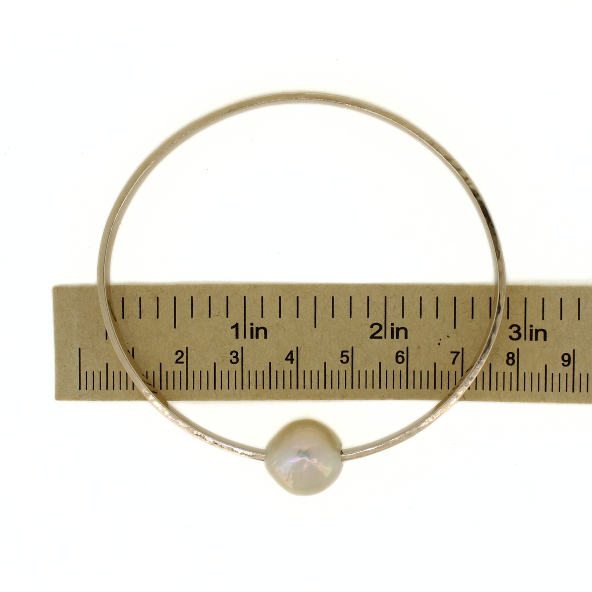 A thick gold oval bangle bracelet with a single round white pearl, shown next to a paper ruler for scale on a white background.