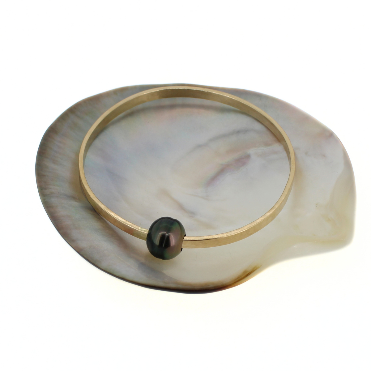Gold hammered bangle bracelet with a dark Tahitian pearl, displayed on a mother of pearl shell