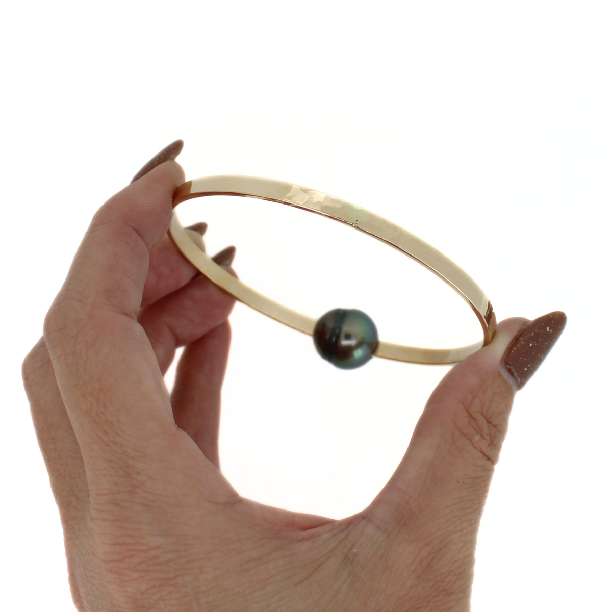 A hand with brown glittery nail polish holds a thick gold bangle bracelet featuring a single dark iridescent pearl, set against a plain white background.