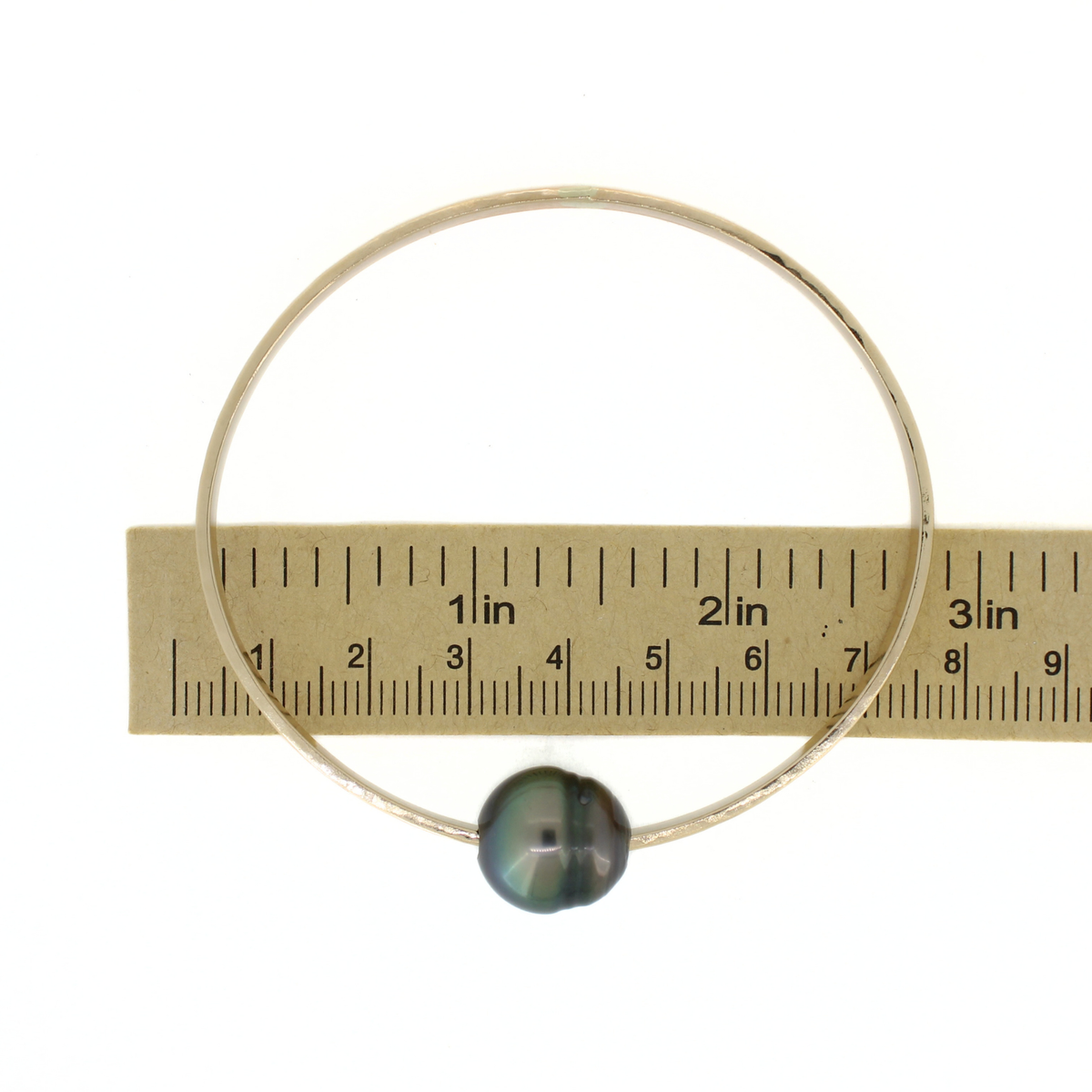 A thick gold bangle bracelet with a single dark green iridescent pearl, shown next to a paper ruler for scale on a white background.