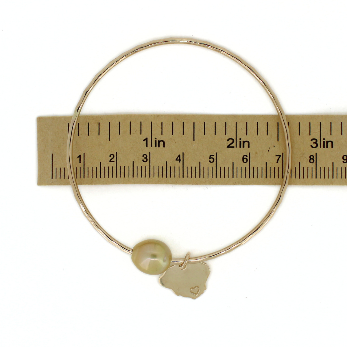 A thin gold bangle bracelet with a single round pearl and a small Kauai-shaped charm, shown against a white background with a paper ruler for scale.