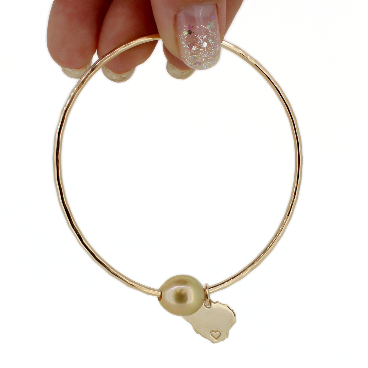 A gold bangle bracelet with a textured finish, featuring a round pearl bead and a small gold Kauai island charm engraved with a heart. The bracelet is held by a hand with glittery nail polish against a solid white background.
