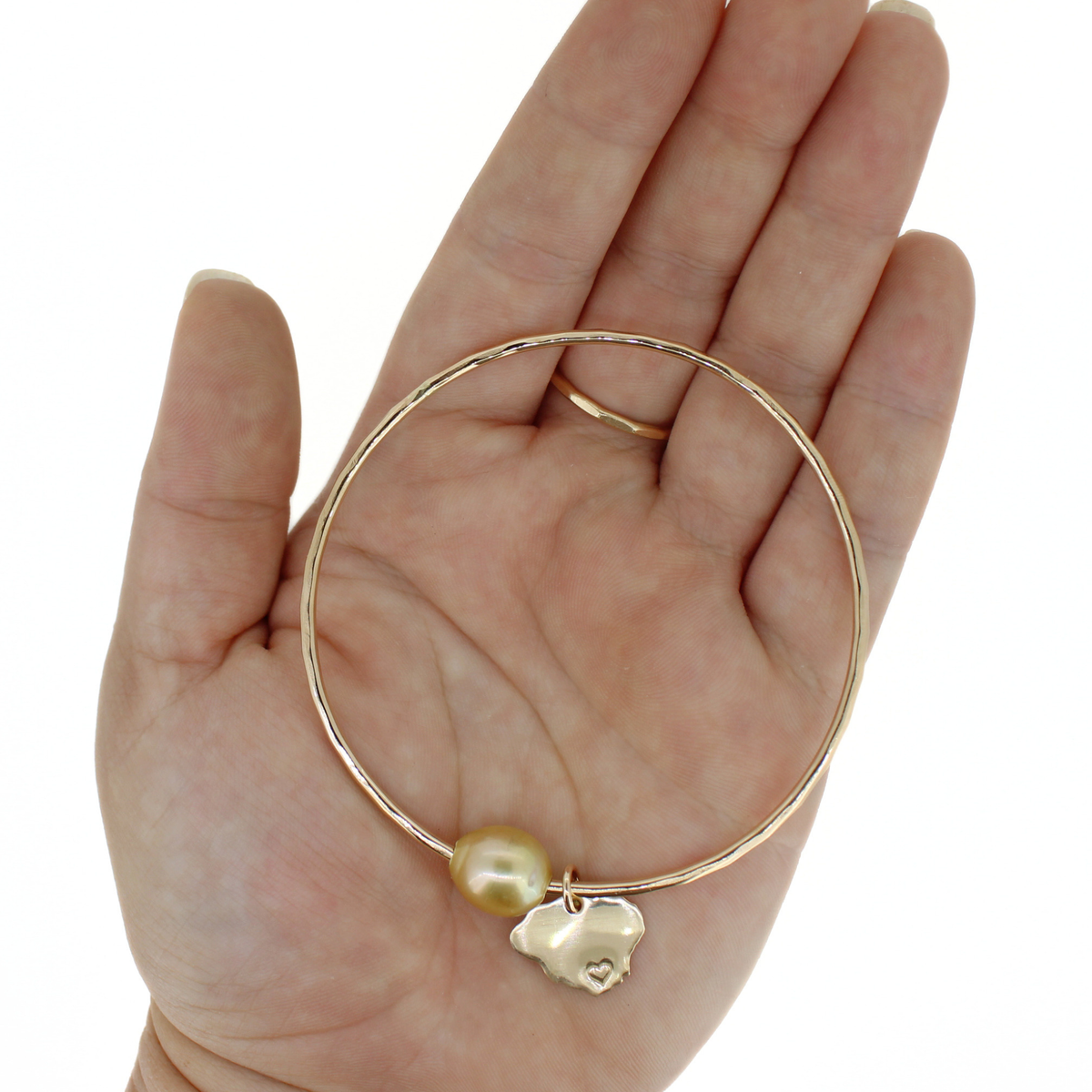 A gold bangle bracelet with a baroque pearl and a small gold Kauai charm, displayed on an open hand against a white background.