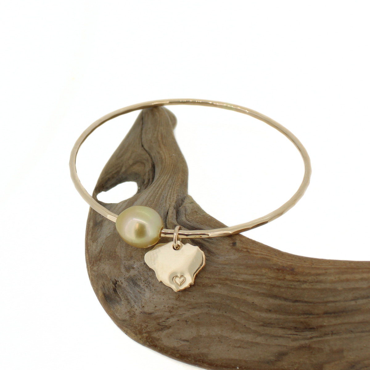 A delicate gold bangle bracelet with a single baroque pearl and a small gold Kauai island charm, displayed on a piece of dark driftwood against a white background.