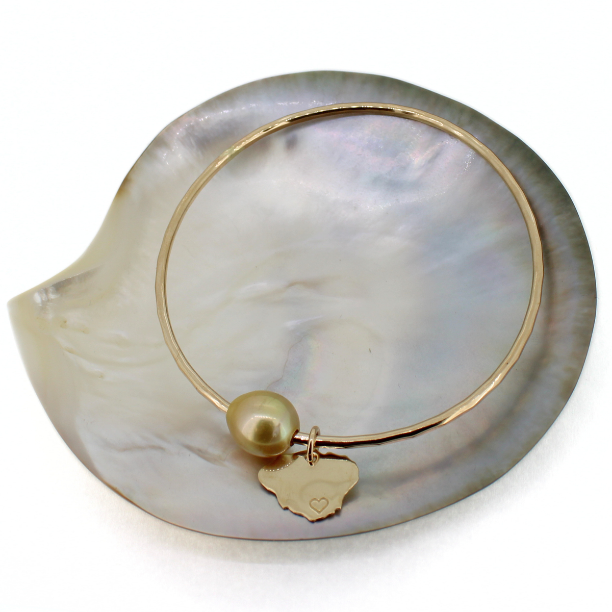 A delicate gold bangle bracelet with a baroque pearl and a small gold kauai island charm engraved with a heart, displayed on a white mother-of-pearl shell.