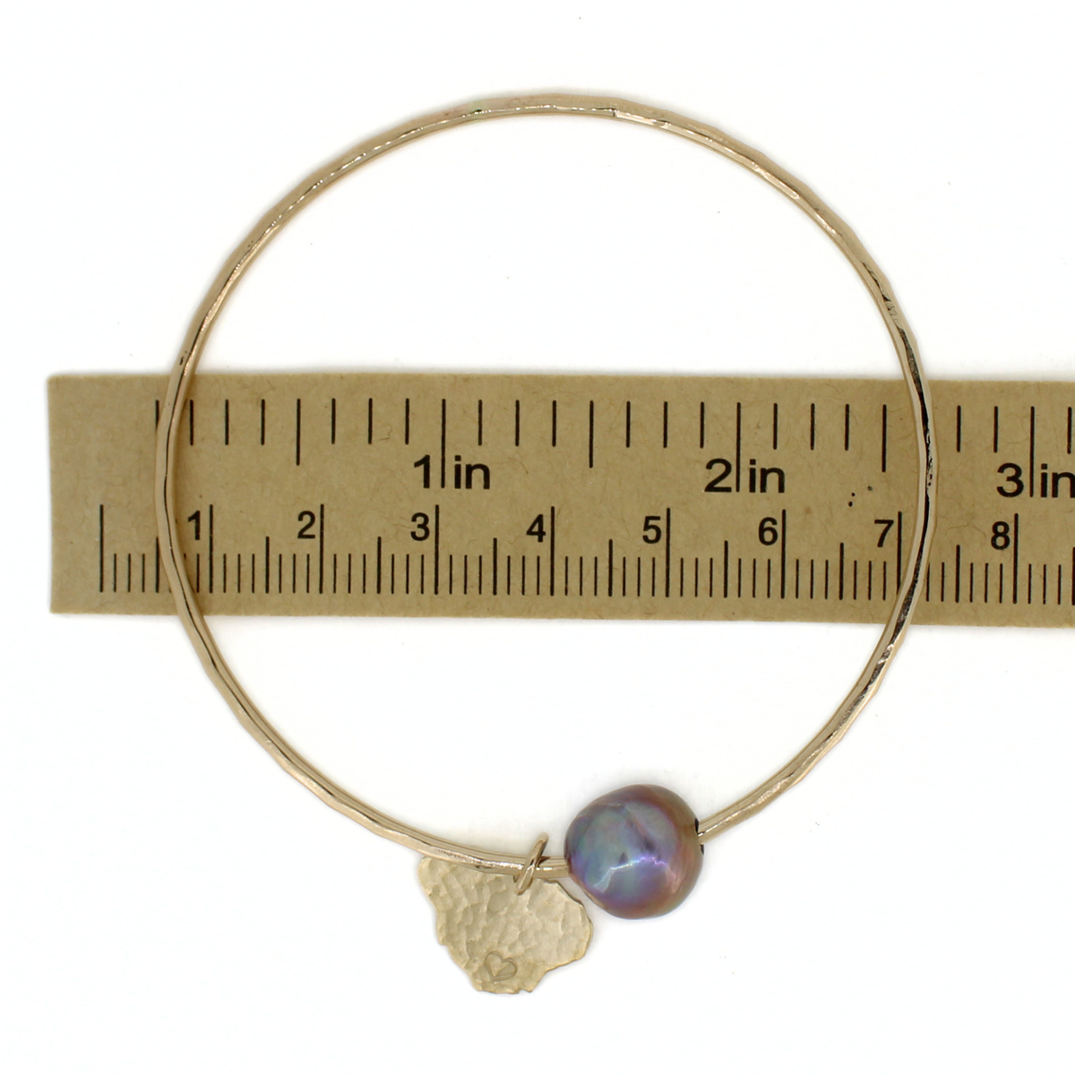 A thin gold bangle bracelet with a small iridescent pearl and a hammered gold charm, shown resting on a paper ruler for scale against a white background.