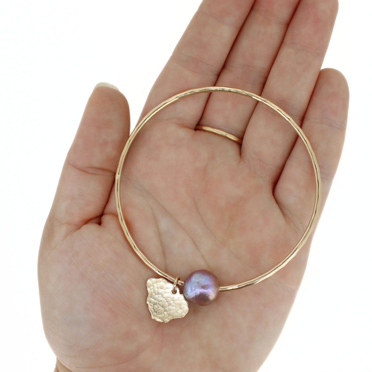 A gold bangle bracelet with a hammered gold Kauai charm and a baroque pearl, held in a person's hand against a white background.