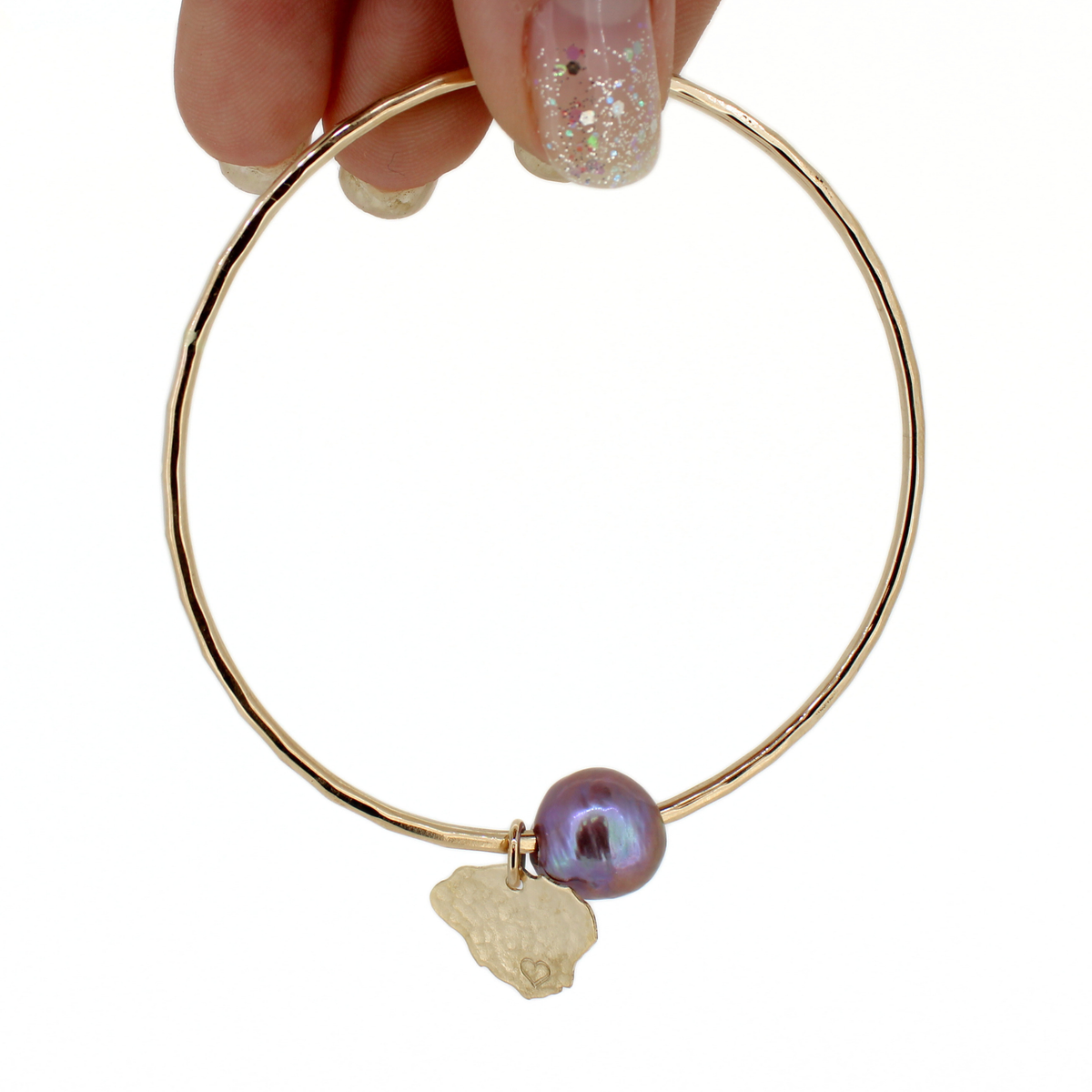 A gold bangle bracelet with a small iridescent purple pearl and a textured gold Kauai charm, held by a hand with glittery nail polish against a white background.