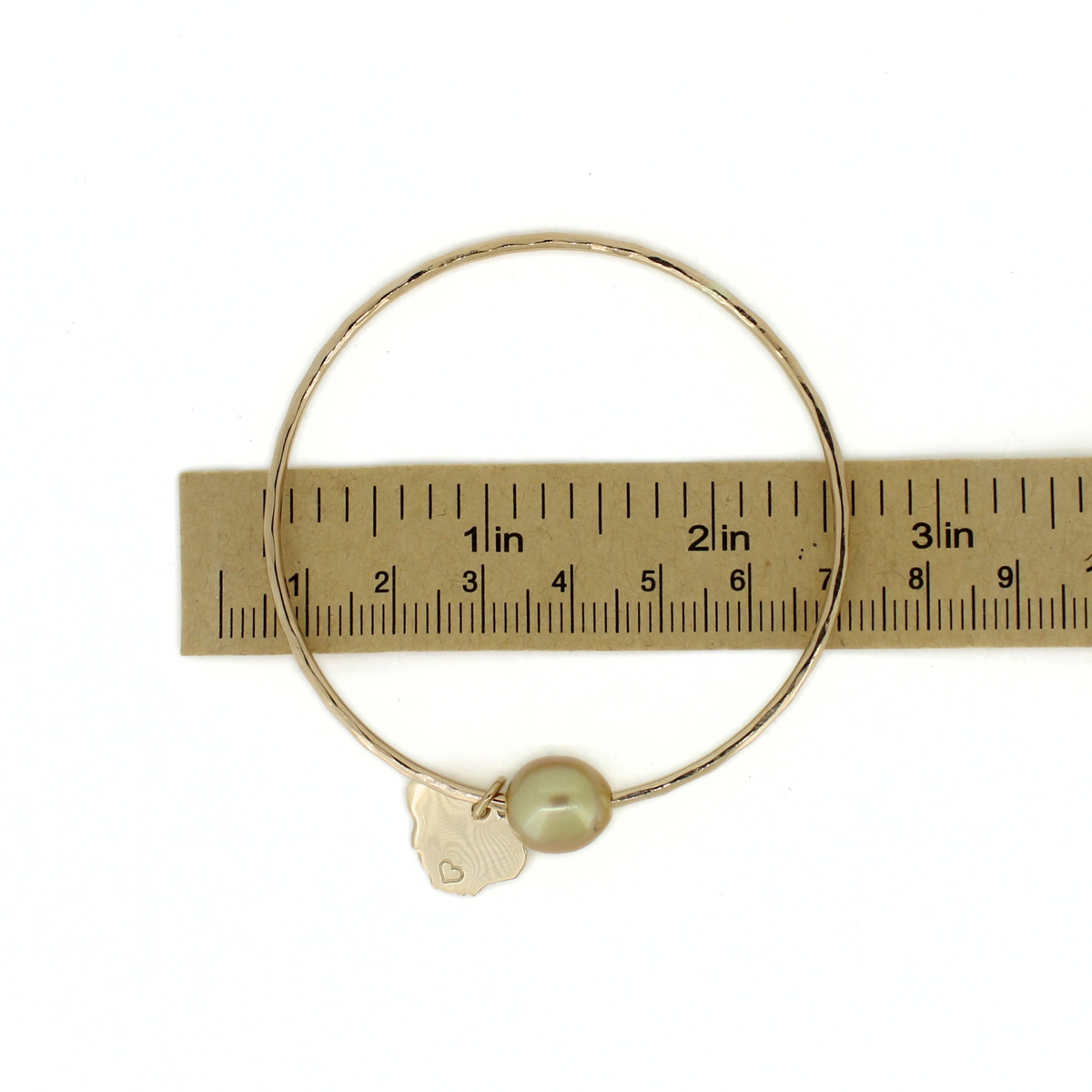 A gold bangle bracelet with a small gold Kauai charm and a single pearl bead, shown next to a paper ruler for scale on a white background.