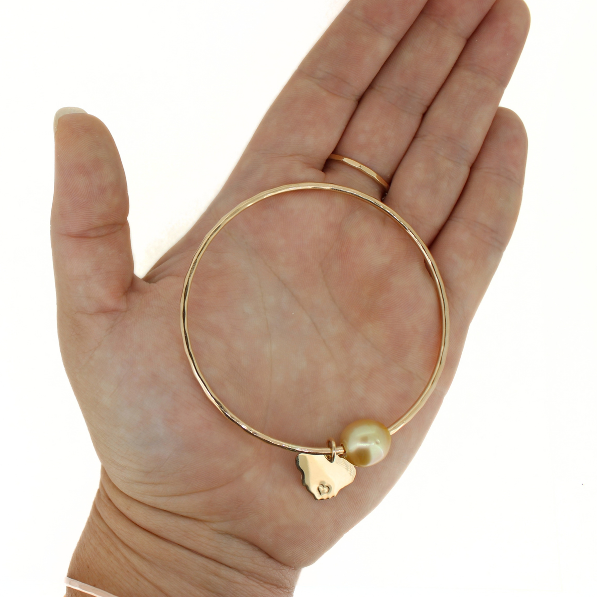A gold bangle bracelet with a small Kauai charm and a pearl bead, displayed on an open hand against a white background.
