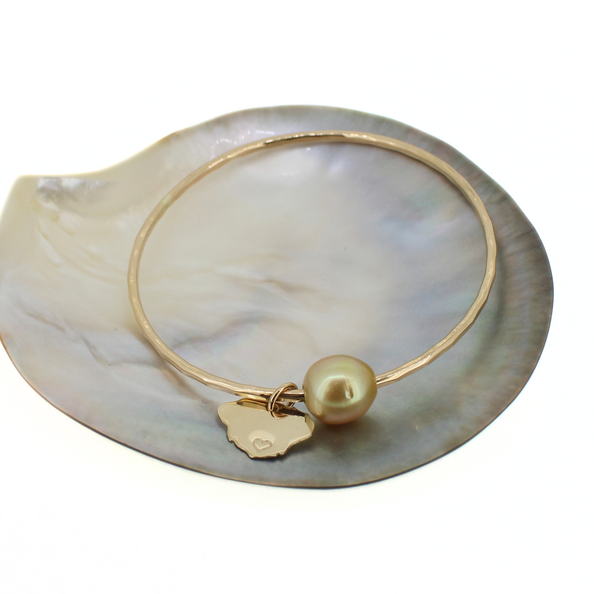 A gold bangle bracelet with a small gold Kauai charm and a round pearl bead, displayed on a white mother-of-pearl shell.