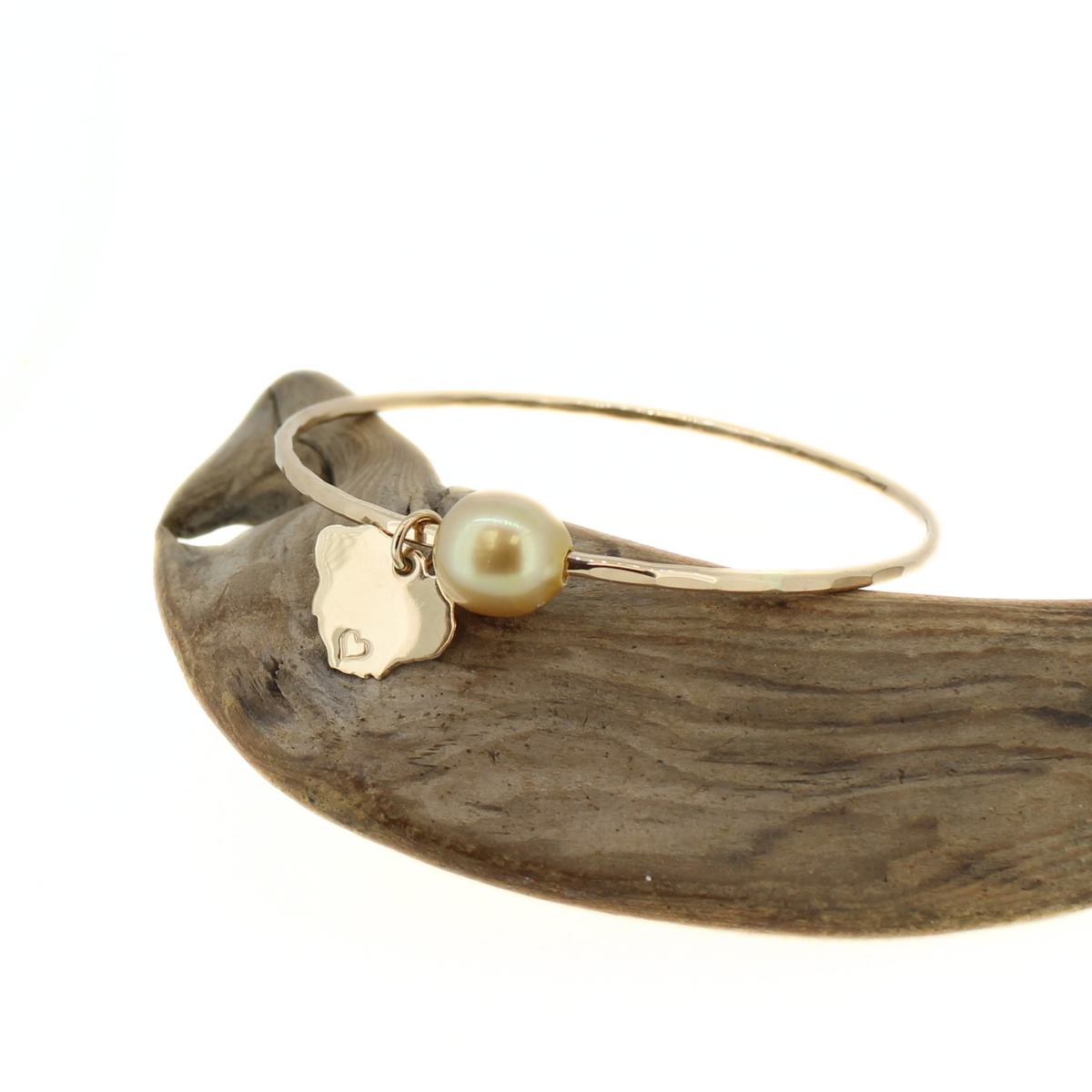 A gold bangle bracelet with a small gold Kauai island charm and a single gold pearl, displayed on a piece of driftwood against a white background.