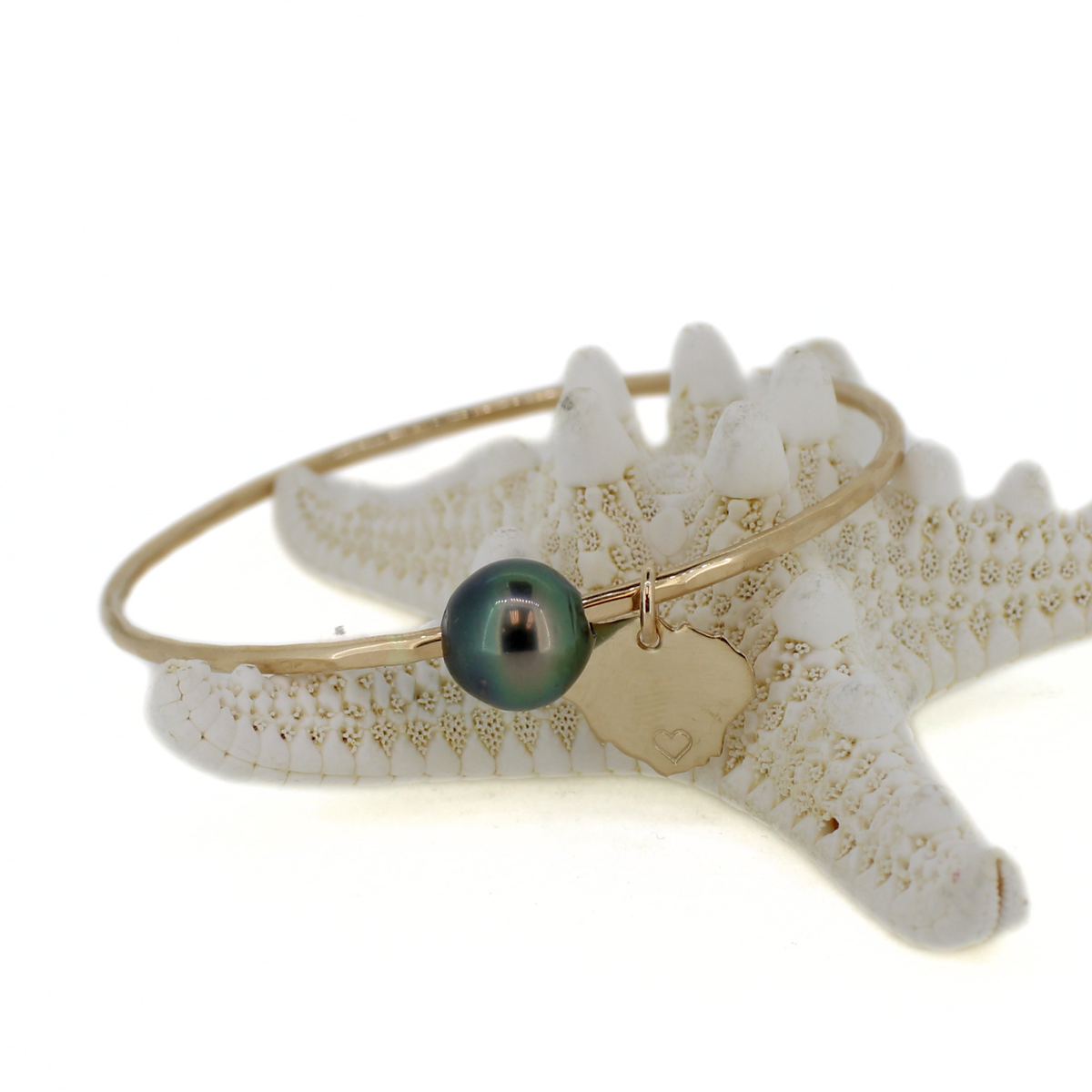 A gold bangle bracelet with a dark green iridescent pearl and a small gold Kauai charm, displayed on a white starfish prop against a white background.