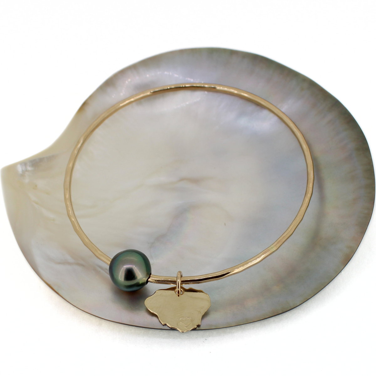 A gold bangle bracelet with a dark green/black pearl and a small gold Kauai charm, displayed on a white mother-of-pearl shell.