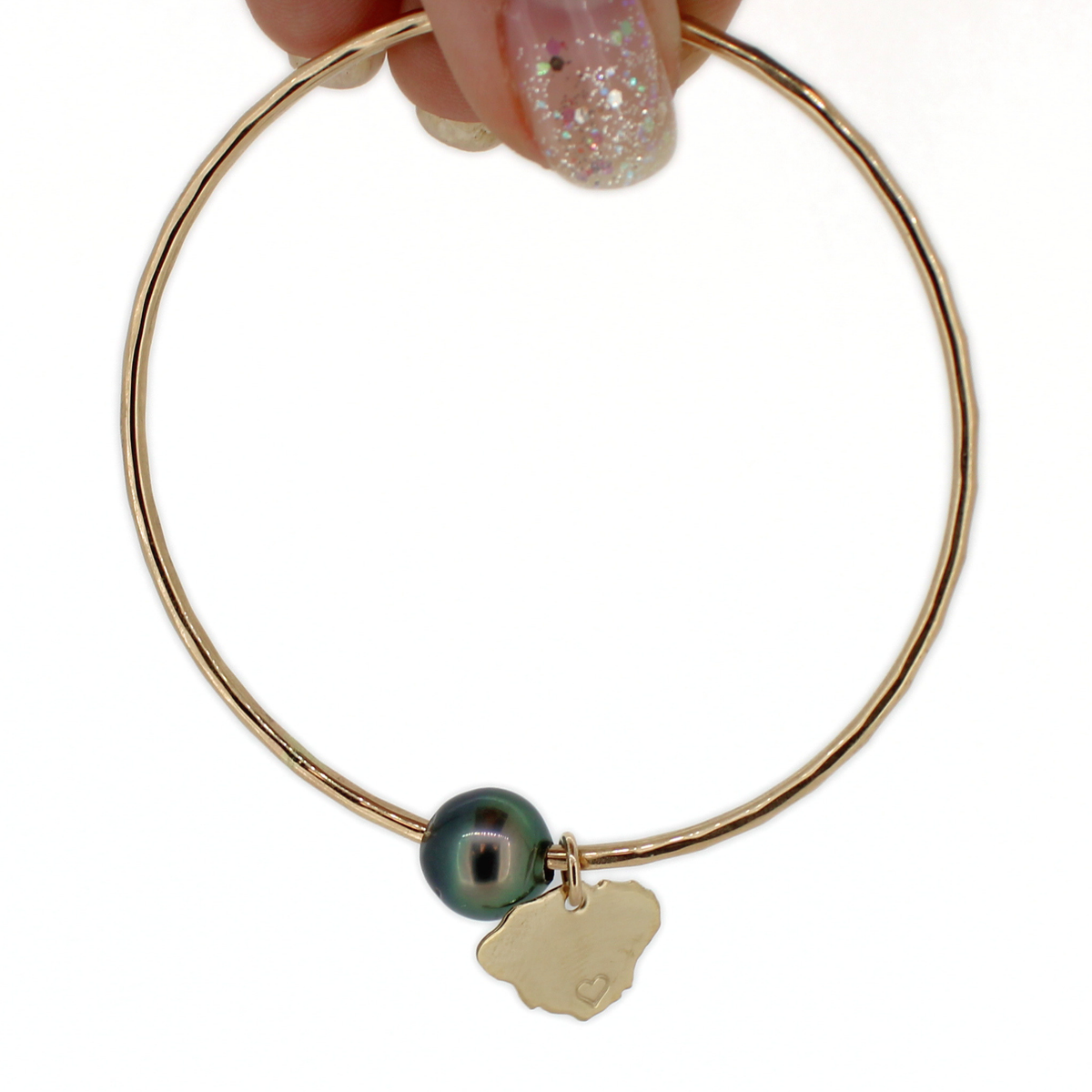 A gold bangle bracelet with a dark green pearl and a small gold Kauai charm, held by a hand with glittery nail polish against a white background.