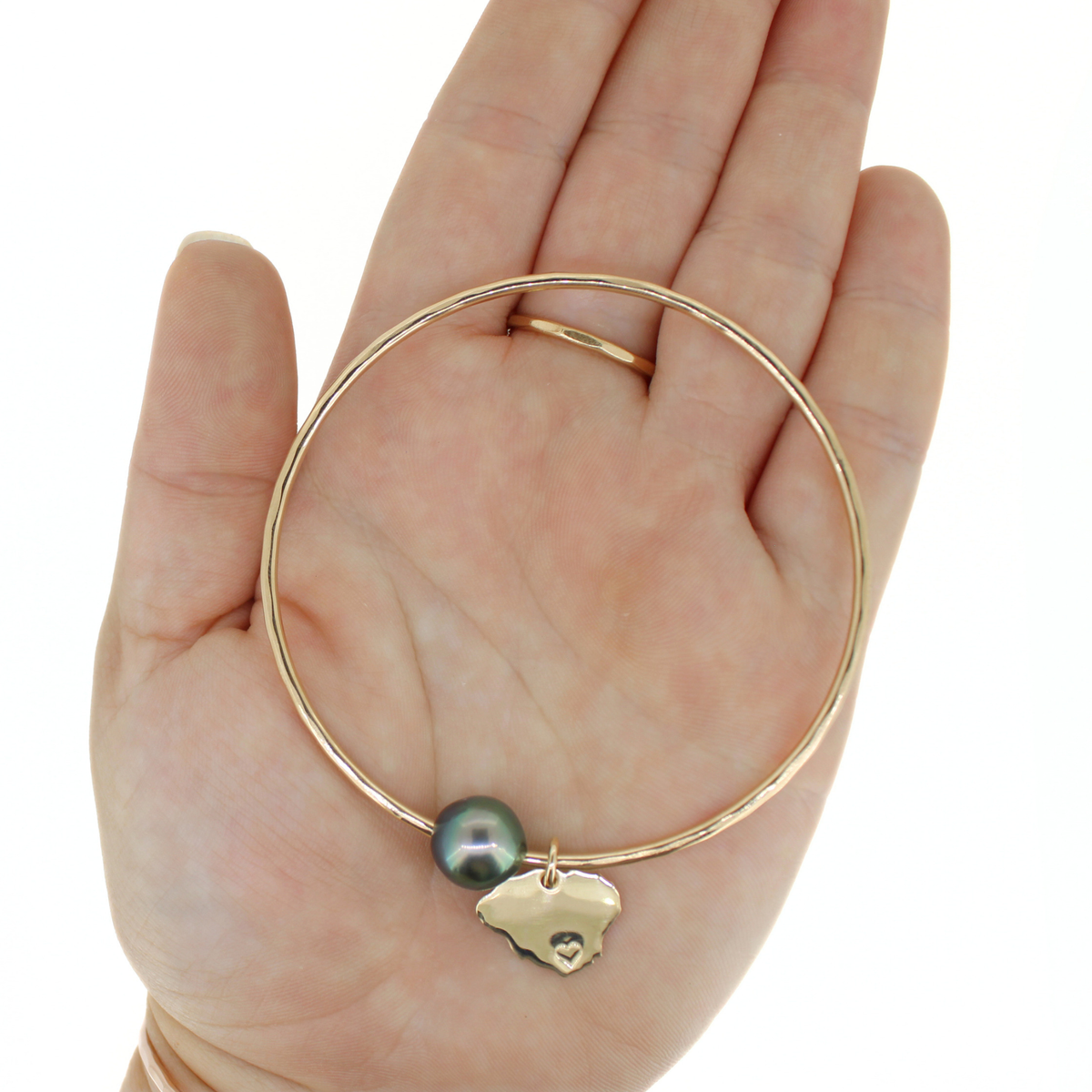 A gold bangle bracelet with a small dark pearl and a gold Kauai charm, displayed on a person's palm.