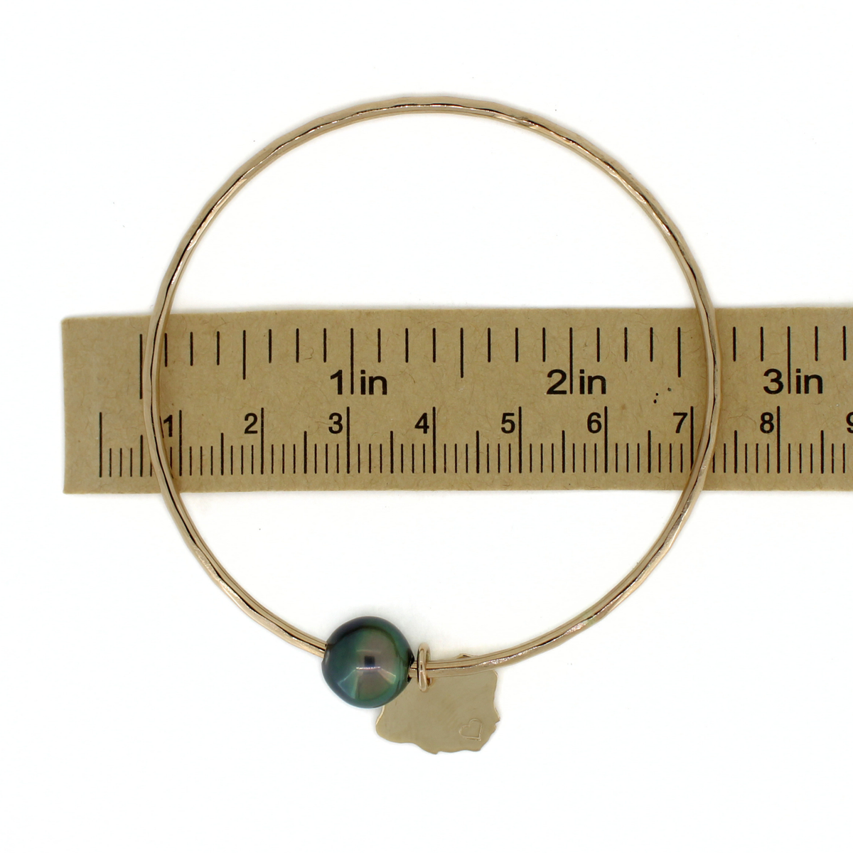 A thin gold bangle bracelet with a dark green Tahitian pearl and a small gold Kauai charm, shown next to a paper ruler for scale on a white background.