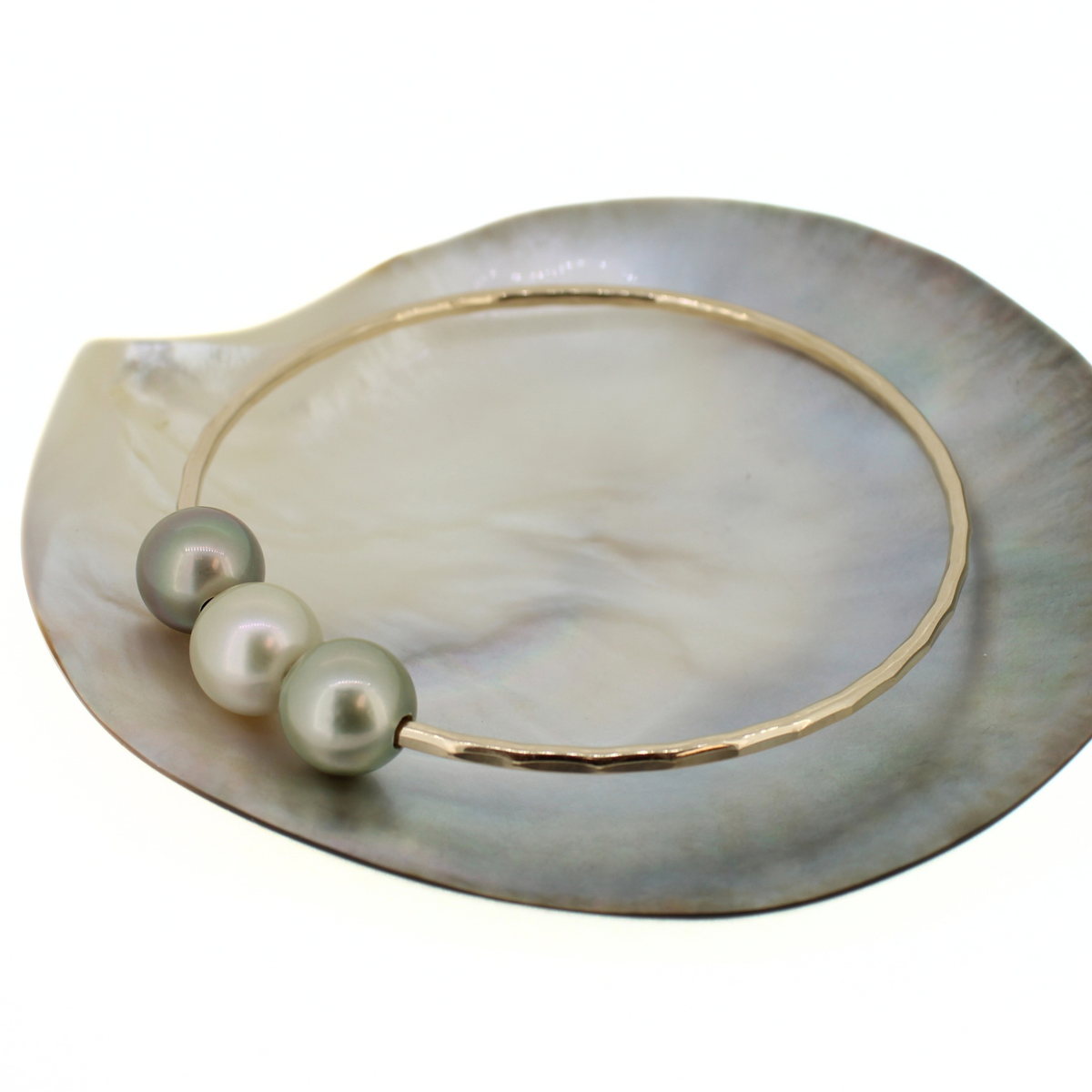 A gold hammered bangle bracelet with three greyish-green Tahitian pearls, displayed on a mother-of-pearl shell dish against a white background.