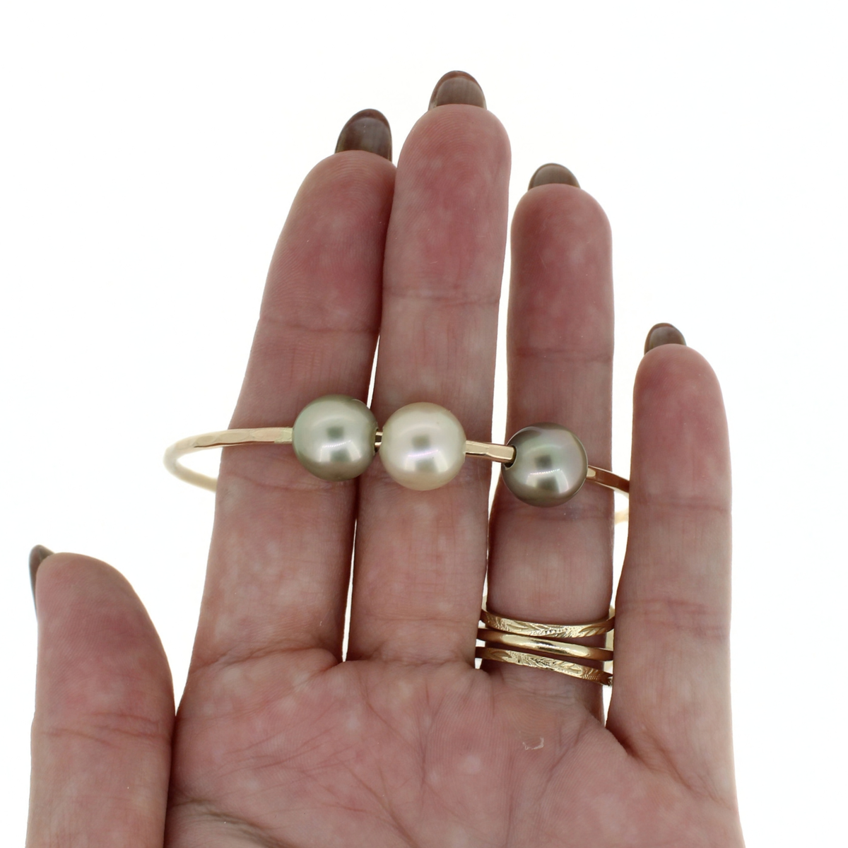 A hand holding a gold bangle bracelet with three Tahitian pearls, against a plain white background.
